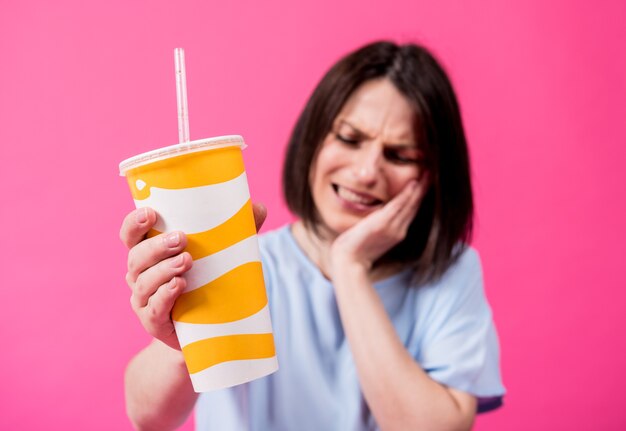 Young woman with sensitive teeth drinking cold water on color background