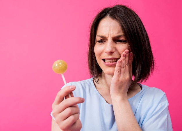 色の背景に甘いロリポップを食べる敏感な歯を持つ若い女性