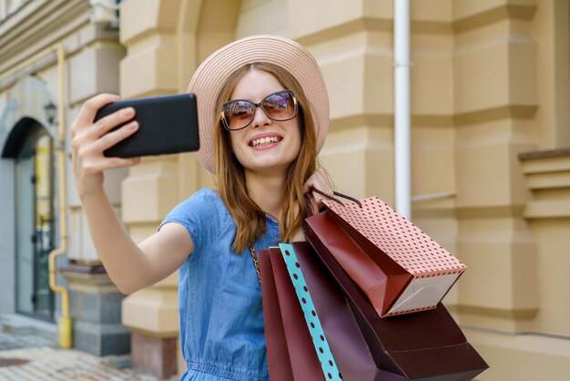 Young woman with shopping bags making selfie walking in a city at summer day