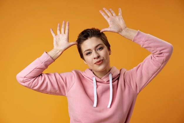 Young woman with short hair doing funny gesture over head as bull horns over yellow background