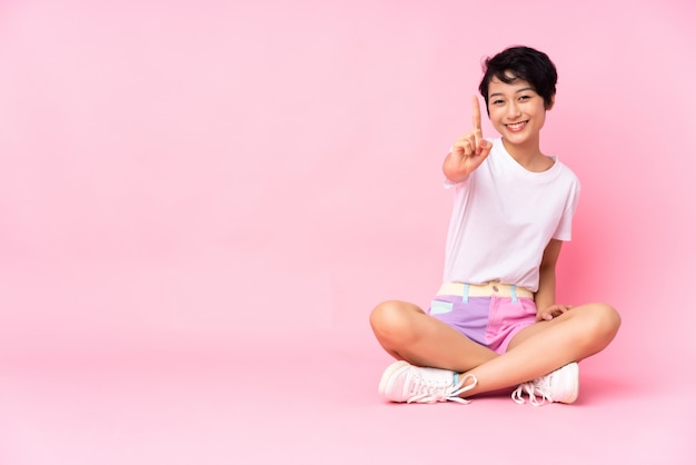 Young woman with short hair sitting on the floor over pink wall showing and lifting a finger