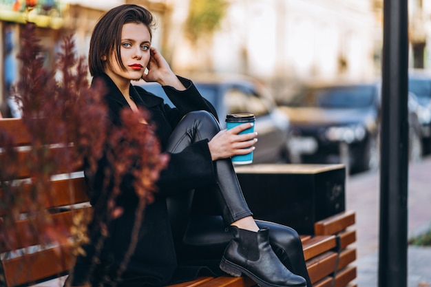 Young woman with short haircut, dressed in black, sits on a bench in the city. A woman holds a cup of coffee