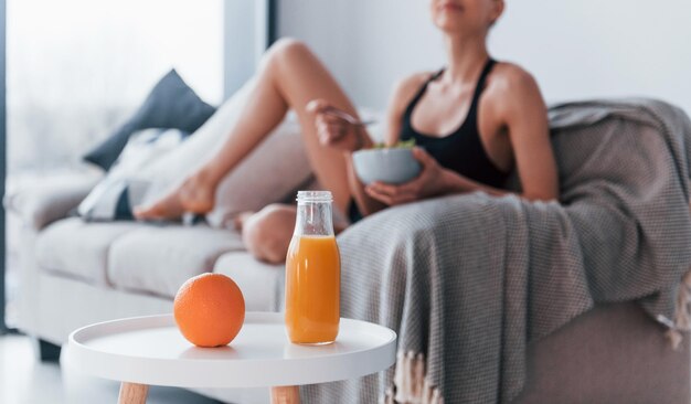 Young woman with slim body shape in sportswear sits on sofa and eats healthy diet food indoors at home