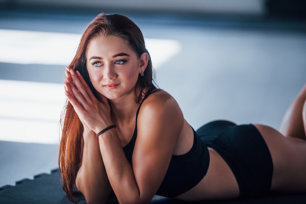 Young woman with slim type of body and in black sportive clothes lying down on the fitness mat.