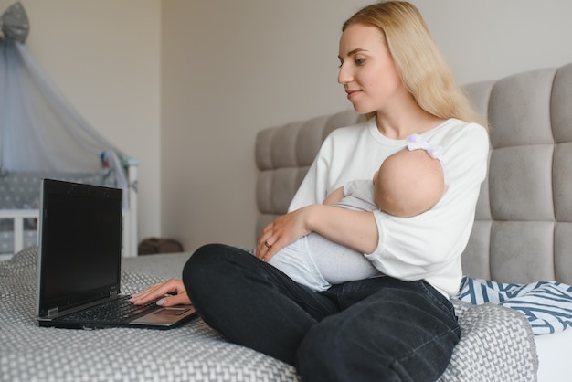 Una giovane donna con un bambino piccolo lavora da casa. lavorare da remoto durante il lockdown. libero professionista o specialista impiegato a distanza è seduto su un letto con un laptop e cerca di concentrarsi