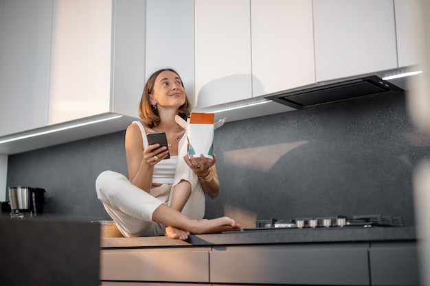 Young woman with smart phone and takeaway food sitting relaxed on the modern kitchen. Concept of ordering food and not cooking at home. Modern lifestyle and living during pandemic at home