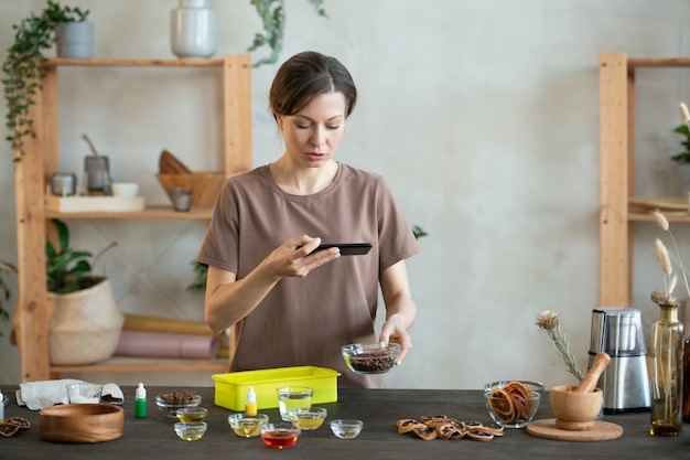 Photo young woman with smartphone shooting video while standing by table and touching small bowl containing essence oil for handmade soap