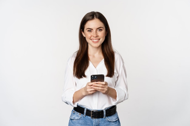 Молодая женщина со смартфоном улыбается и смотрит в камеру, используя сотовую технологию приложения для мобильного телефона...