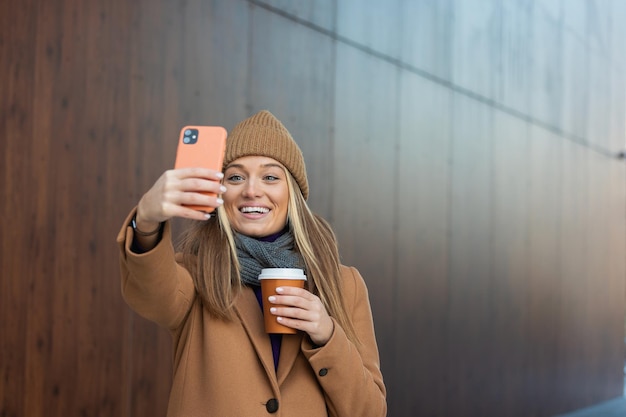 Young woman with smartphone standing against street blurred building background Fashion photo of beautiful girl with phone and cup of coffee