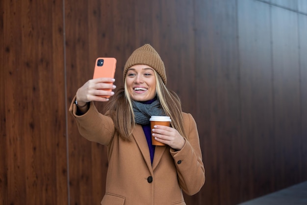 Young woman with smartphone standing against street blurred building background Fashion photo of beautiful girl with phone and cup of coffee