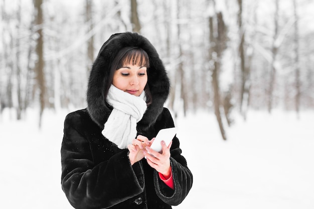 Young woman with smartphone and winter landscape snowflakes on the background.