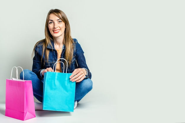 Young woman with a smile in a denim jacket holds shopping bags while sitting on the floor on a light space. Banner. Concept shopping.