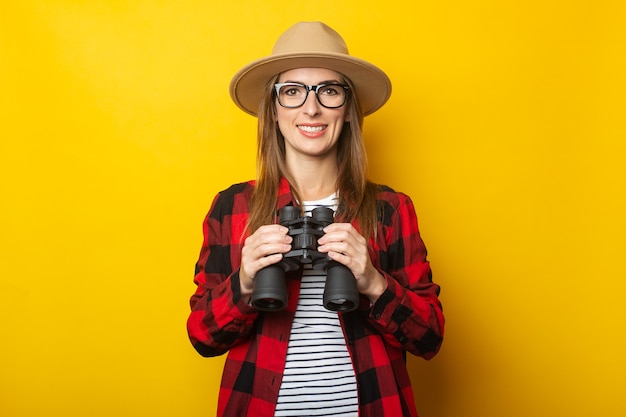 Young woman with a smile in a hat and a plaid shirt holding binoculars in her hands on a yellow background.