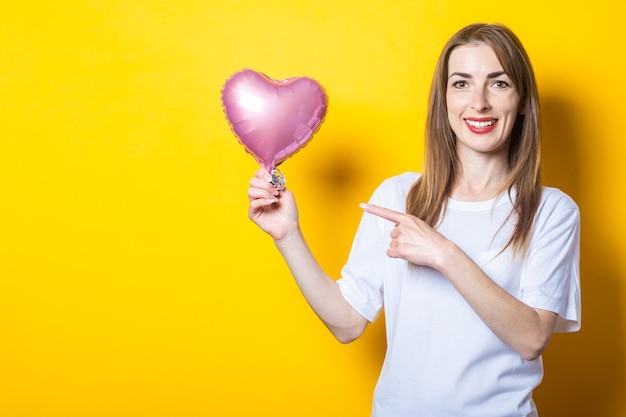 Young woman with a smile holds a heart-shaped balloon in her hands and points her finger at it on a yellow background. Banner.