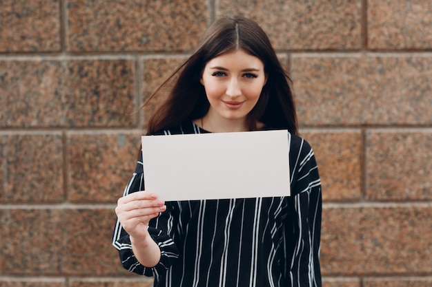 Young woman with smile holds white paper in hand stone background. Girl with white blank template sheet with empty space.