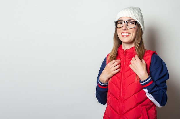 Young woman with a smile in a red vest, hat and glasses against a light wall