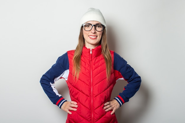 Photo young woman with a smile in a red vest, hat and glasses against a light wall