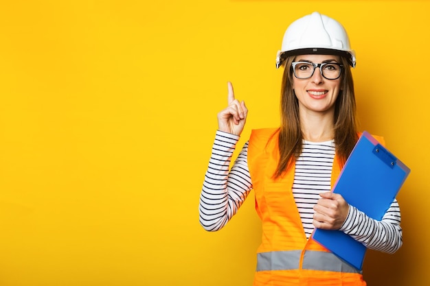 ベストとヘルメットの笑顔で若い女性は黄色の背景にクリップボードを保持します建設コンセプト新築バナー