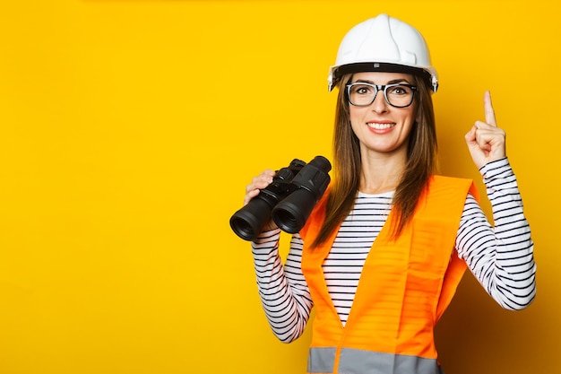 Young woman with a smile in a vest and a helmet holds binoculars on a yellow background construction concept new building banner