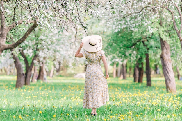 麦わら帽子を持つ若い女性は、春に花の咲く庭園を歩きます。桜の木の下の女の子。夏のファッションのコンセプトです。