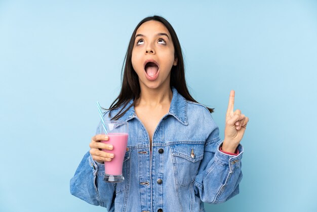 Young woman with strawberry milkshake over blue surprised and pointing up