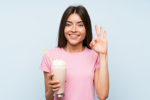 Молодая женщина с клубничный молочный коктейль на изолированных синем фоне, показывая знак ОК с пальцами