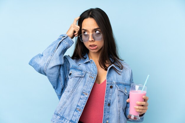 Молодая женщина с клубничным молочным коктейлем на изолированном синем, сомневаясь и думая