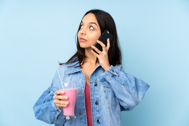 Young woman with strawberry milkshake on isolated blue holding coffee to take away and a mobile