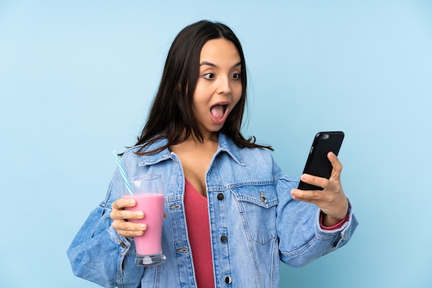 Young woman with strawberry milkshake on isolated blue holding coffee to take away and a mobile