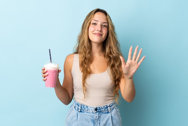 Young woman with strawberry milkshake isolated on blue wall counting five with fingers