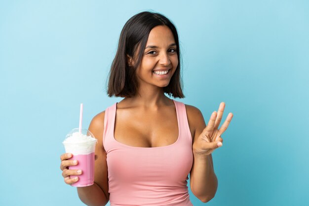 Young woman with strawberry milkshake isolated on blue wall happy and counting three with fingers