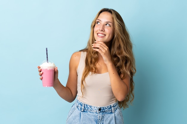 Young woman with strawberry milkshake isolated on blue wall looking up while smiling