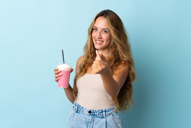 Giovane donna con frappè alla fragola isolato sulla parete blu che fa gesto di soldi