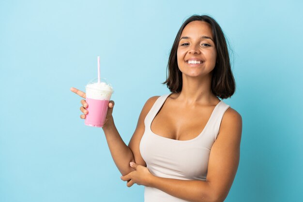 Young woman with strawberry milkshake isolated on blue wall pointing finger to the side