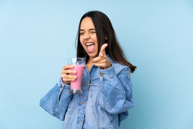 Молодая женщина с клубничным молочным коктейлем над изолированной синей стеной, указывая вперед и улыбаясь
