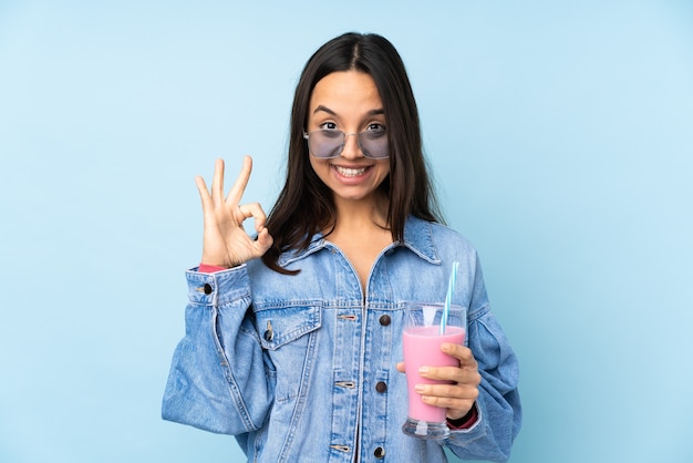 Young woman with strawberry milkshake over isolated blue wall showing ok sign with fingers