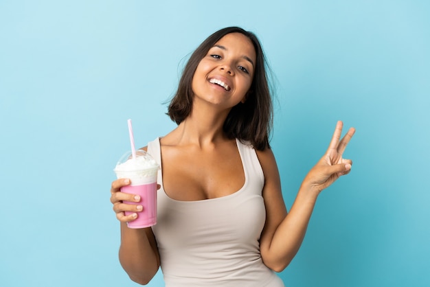 Young woman with strawberry milkshake isolated on blue wall smiling and showing victory sign