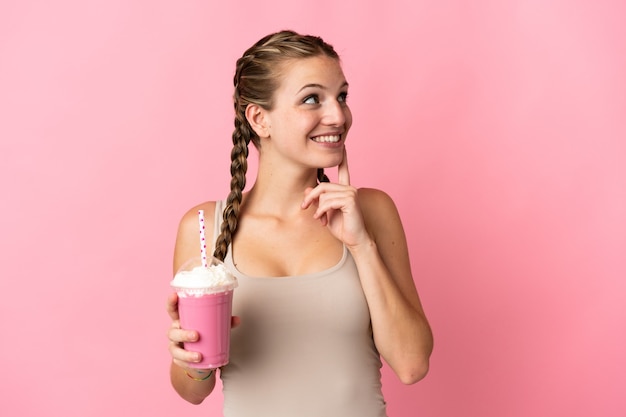 Молодая женщина с клубничным молочным коктейлем изолирована на розовой стене, думая об идее, глядя вверх
