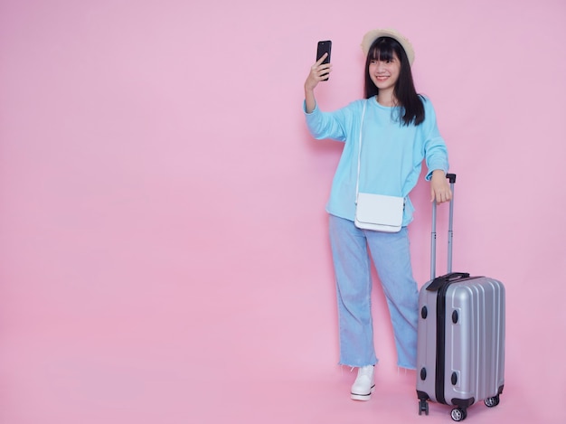 ピンクの壁にスーツケースとスマートフォンを持つ若い女性