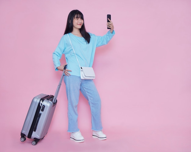 Young woman with suitcase and smartphone on pink wall