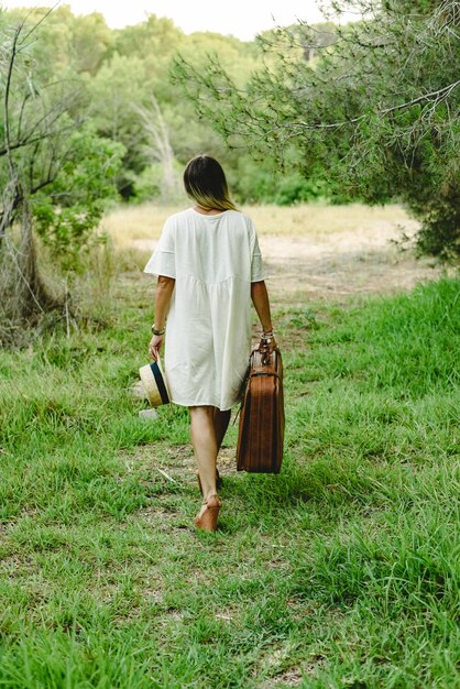 Young woman with suitcase and straw hat in the woods walking\
alone and sad.