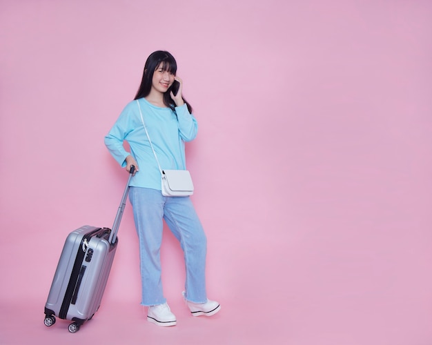 Young woman with suitcase and talking on phone on pink wall