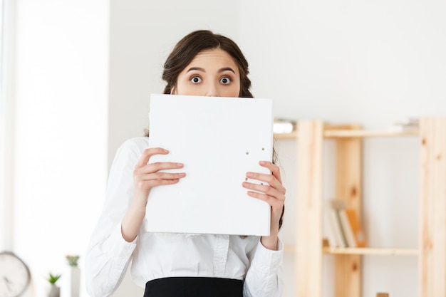 Young woman with surprised eyes peeking out from behind paper poster. Businesswoman holding big white banner in modern office.