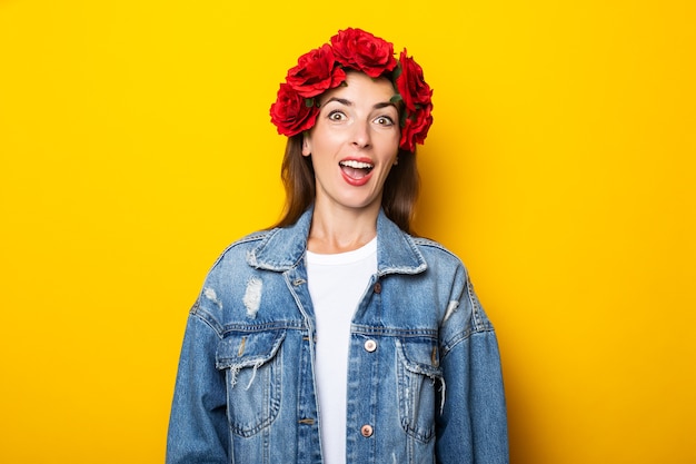 Young woman with a surprised face in a denim jacket and a wreath of red flowers on her head on a yellow wall.