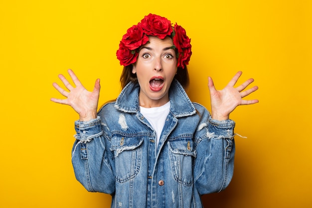 Young woman with a surprised face in a denim jacket and a wreath of red flowers on her head on a yellow wall.