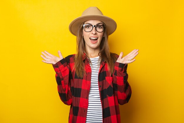 Young woman with surprised face in hat and plaid shirt on yellow.