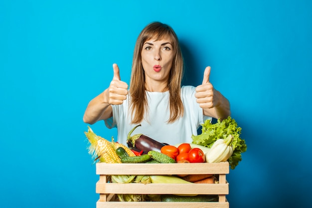 Young woman with a surprised face holds a box with fresh vegetables on blue. good harvest concept, natural product