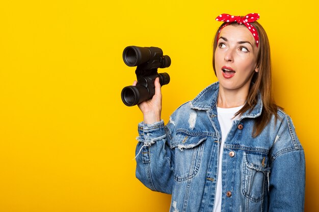 Young woman with a surprised face in jeans and a raft on her head holds binoculars in her hands on a yellow wall. Banner.