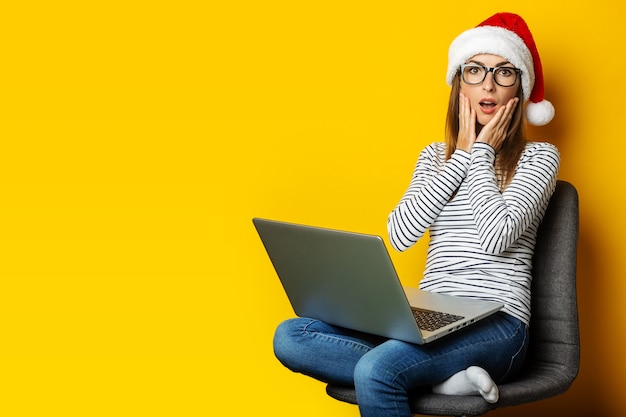 Young woman with a surprised face and a laptop, wearing a santa hat and sitting on a chair