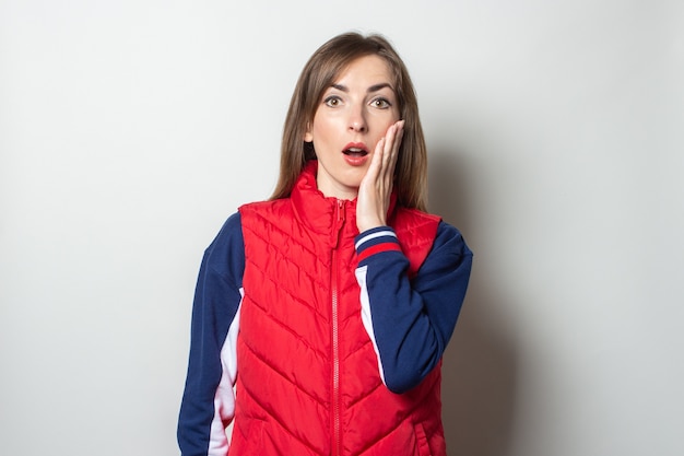 Young woman with a surprised face in a red vest against a light wall